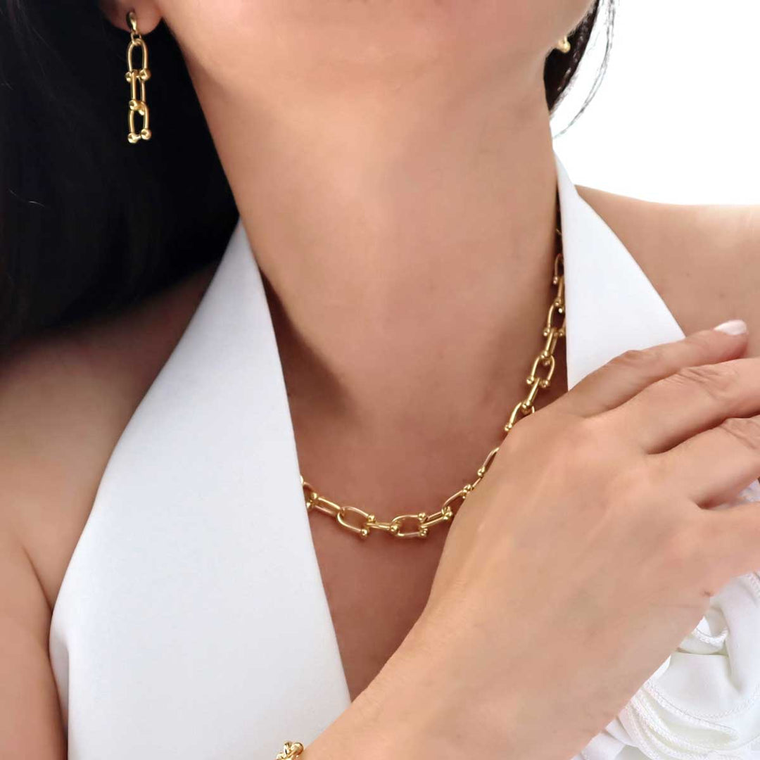 A woman in a white waistcoat wearing a chunky yellow gold gauge link chain necklace with matching bracelet and long dangling gauge link earrings.