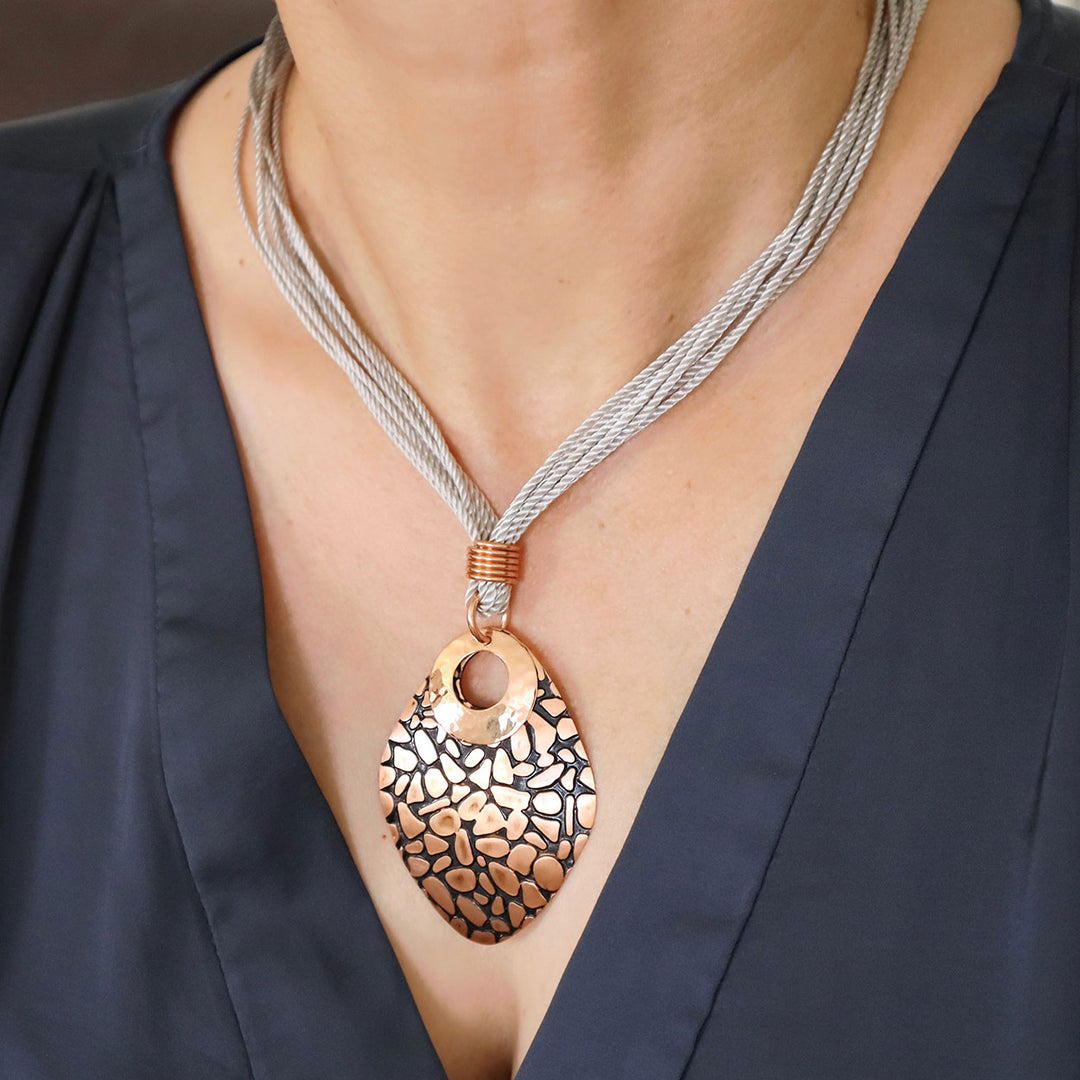 Large polished and brushed copper pendant and cord necklace worn by a woman in a blue silk top