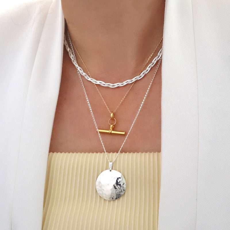 A necklace stack featuring a gold t-bar necklace, a long circle pendant and chain and a braided choker style chain worn by a woman in a white jacket