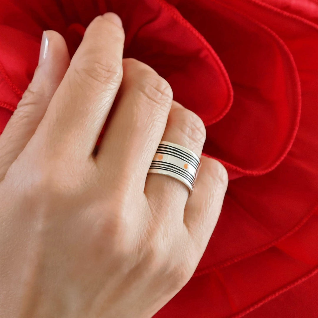 A hand with a wide band silver ring with black lines and copper spots on a red background.