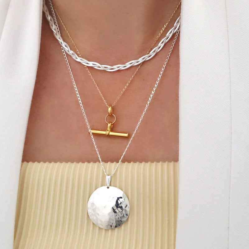 A necklace stack featuring a woven flat silver chain, a gold T-bar and curb chain and a large hammered silver pendant and chain necklace worn by a women with a white jacket.