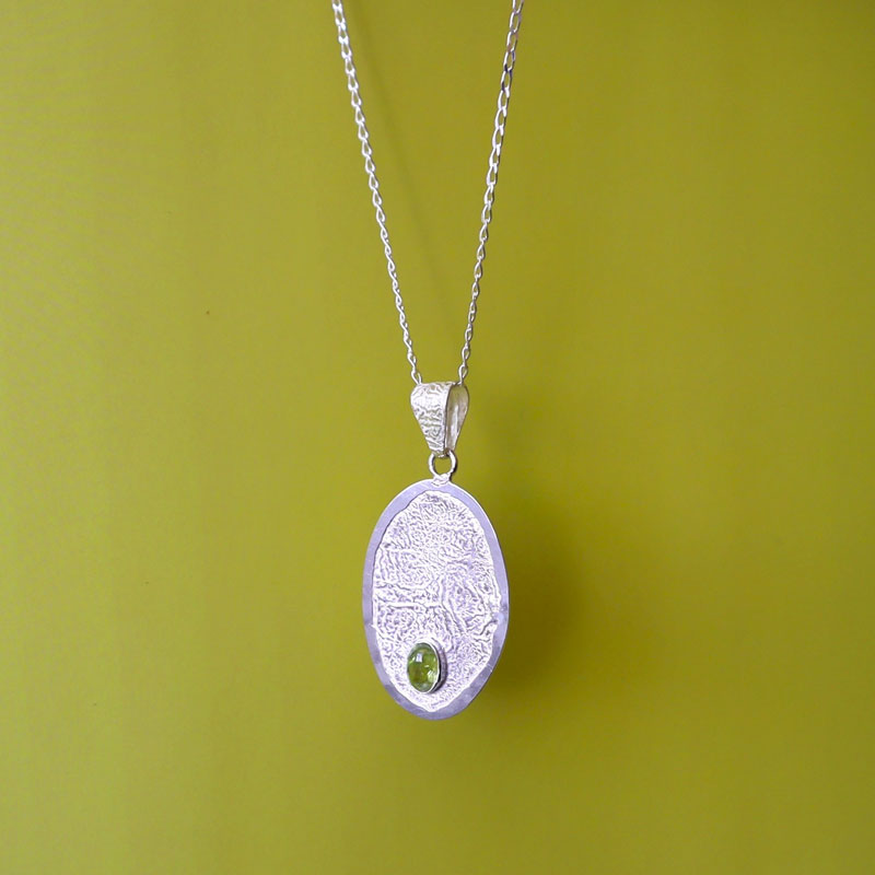 A large unusual, reticulated sterling 925 silver oval handmade pendant and chain necklace decorated with a single peridot gemstone against a green background. 