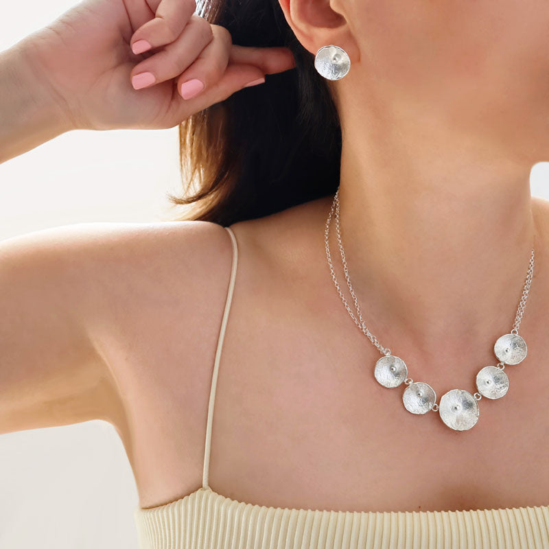 Sterling silver hammered poppy flower necklace and matching stud earrings worn by a woman in a yellow top. 