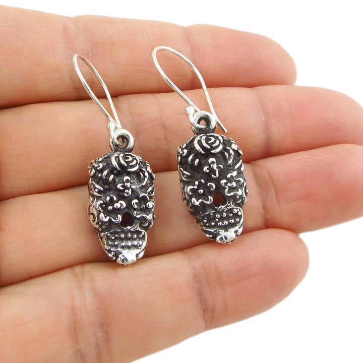 Silver skull-shaped day of the dead calavera earrings held between fingers on a white background