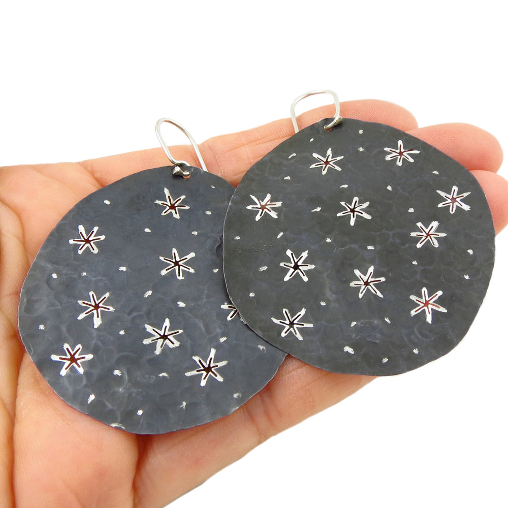 Round silver earrings with a black finish and a star pattern held in a hand against a white background