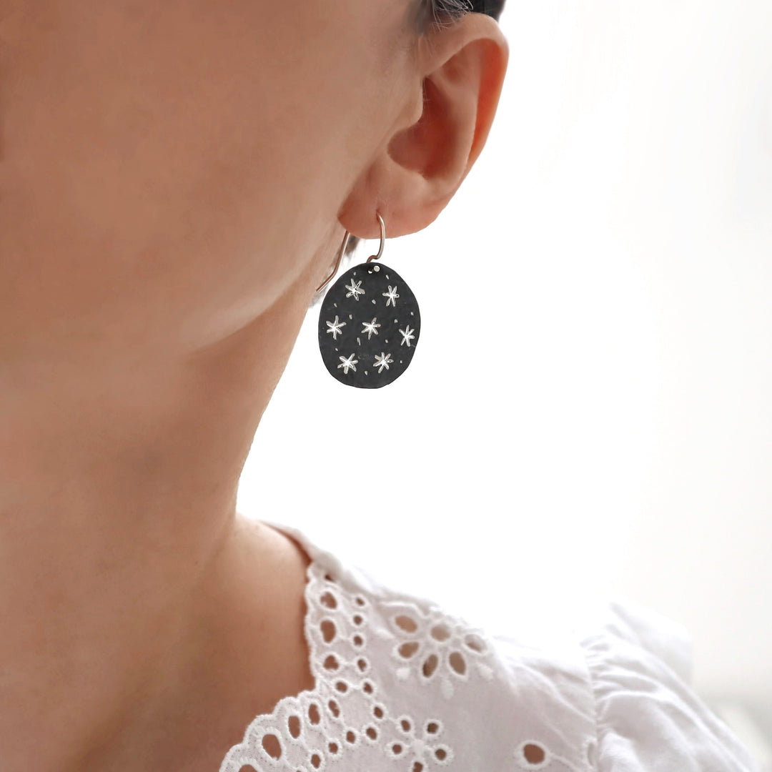 Silver earrings with a black finish and star patterns worn by a person against a white background