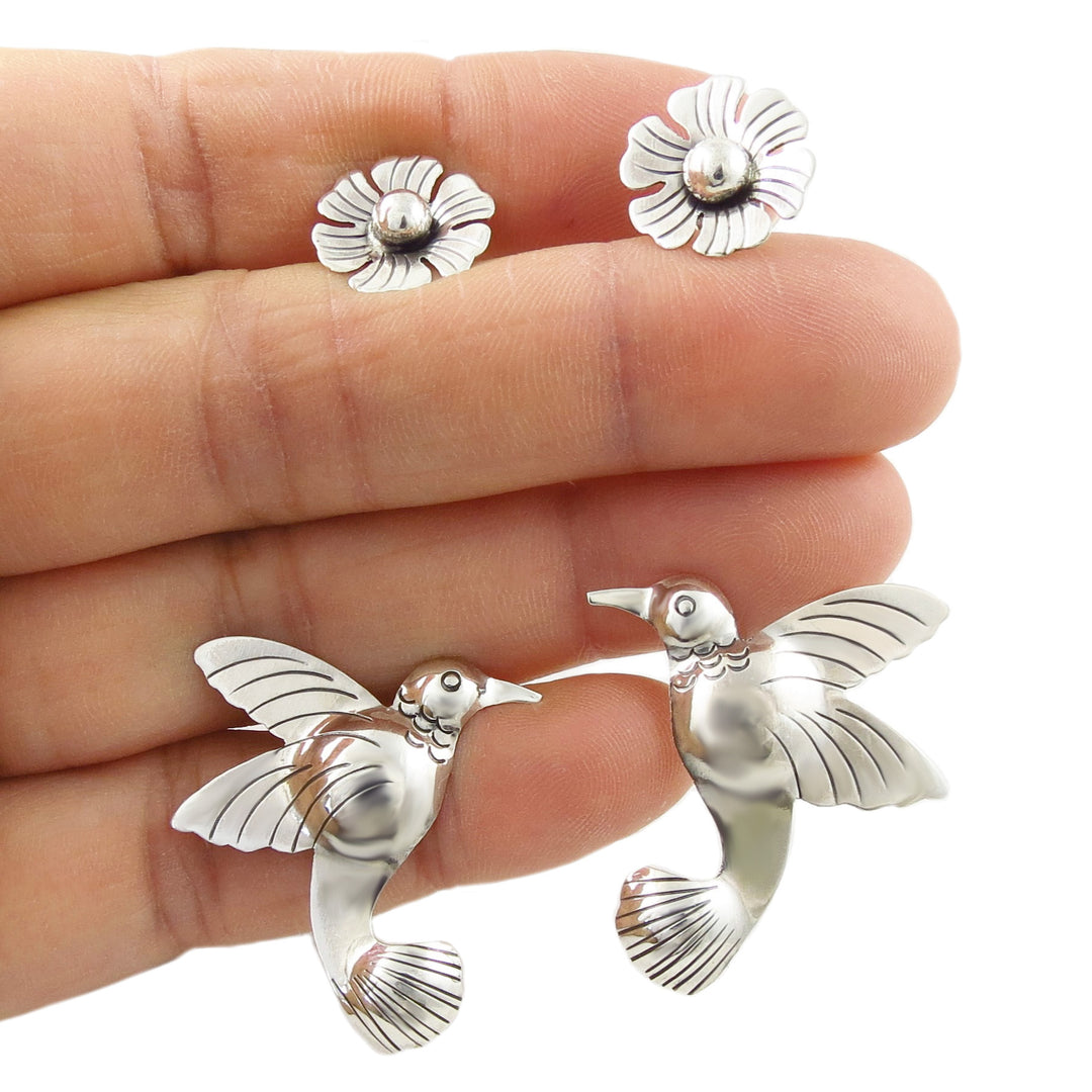 Large sterling silver hummingbird and flower earrings held between fingers on a white background