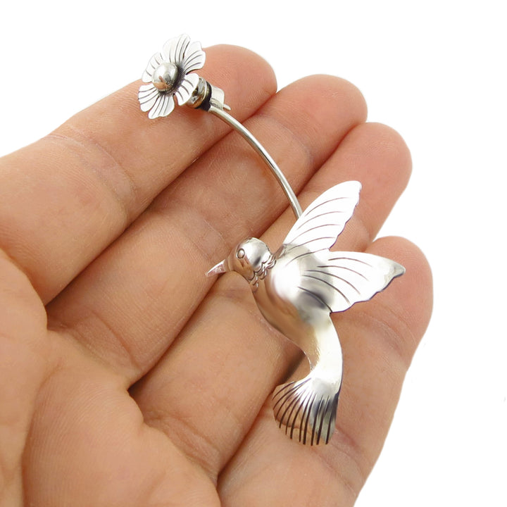 A single Taxco sterling silver hummingbird and flower design earring held in a woman's hand against a white background