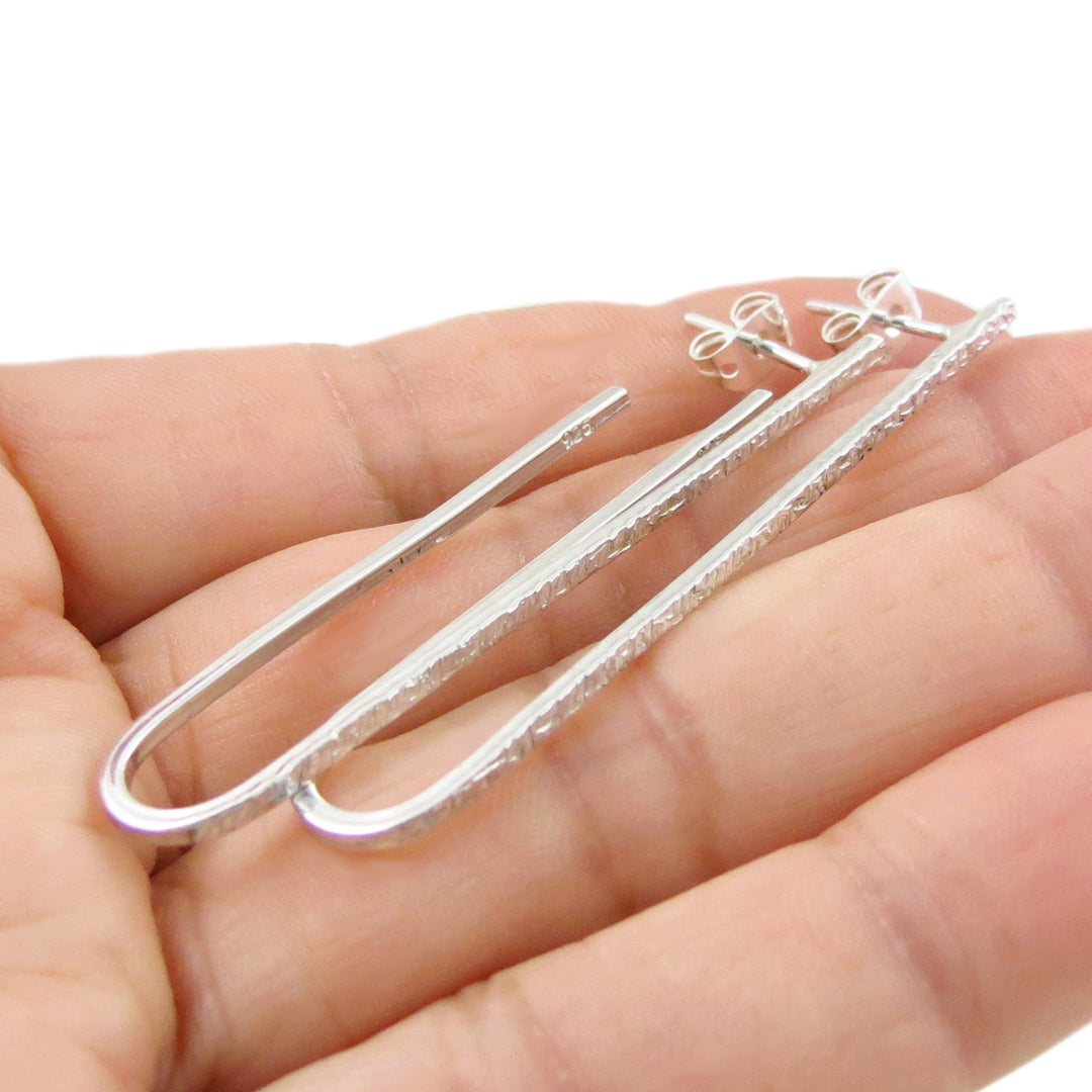A pair of handmade unique 925 sterling silver long oval drop earrings with a battered finish on the front held in a person's hand. 