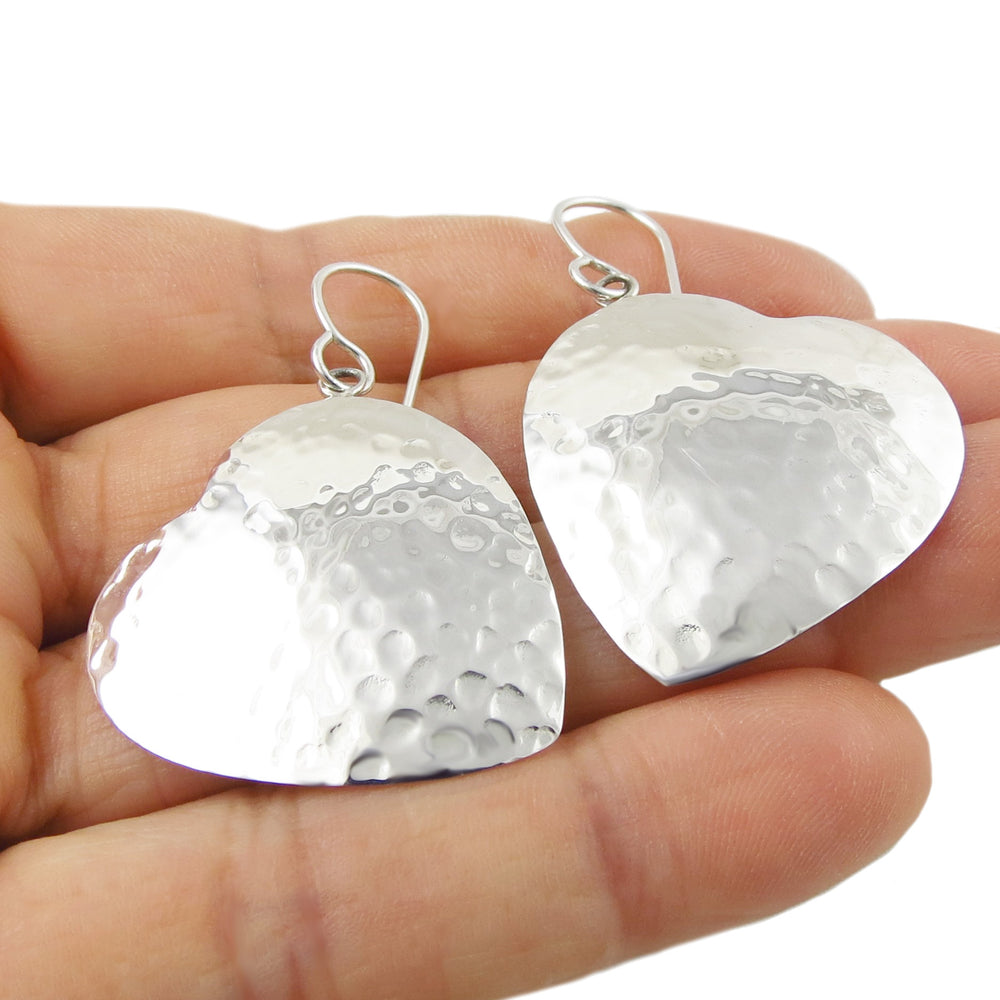 Large silver heart-shaped earrings held in a hand against a white background