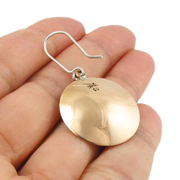 The reverse of a copper disc earring with silver hooks held in a hand with a white background