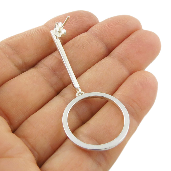 A single statement 925 Silver circle and stick drop earring held between fingers on a white background