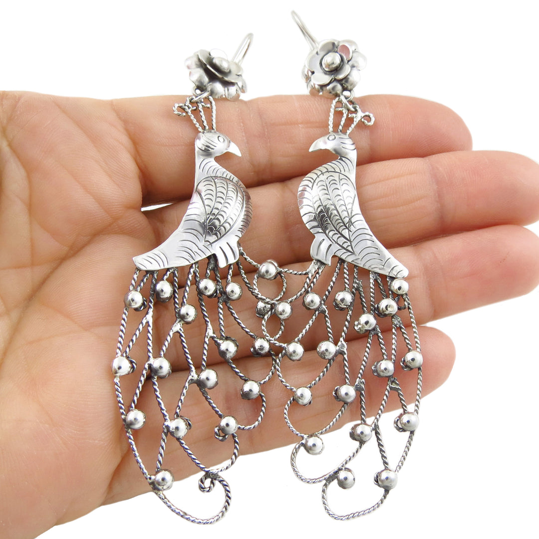 A pair of unique and unusual Mexican silver chandelier earrings featuring long peacock birds with wide tails decorated with rope and bead work held in a person's hand. 