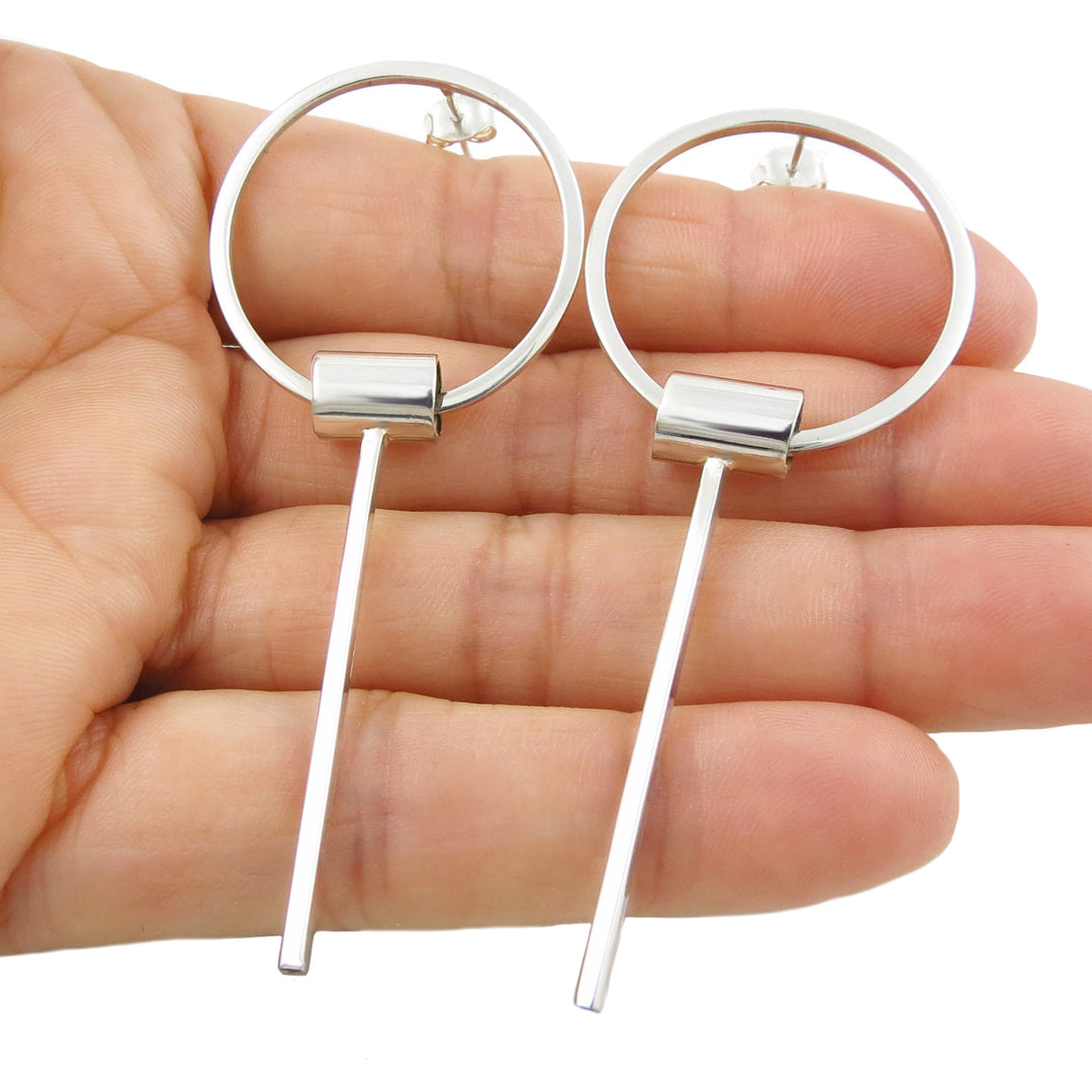 A pair of large 925 sterling silver geometric hoop drop earrings for women held in a person's hand against a white background.
