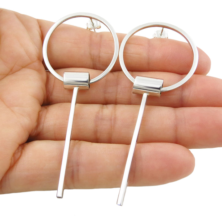 A pair of large 925 sterling silver geometric hoop drop earrings for women held in a person's hand against a white background.