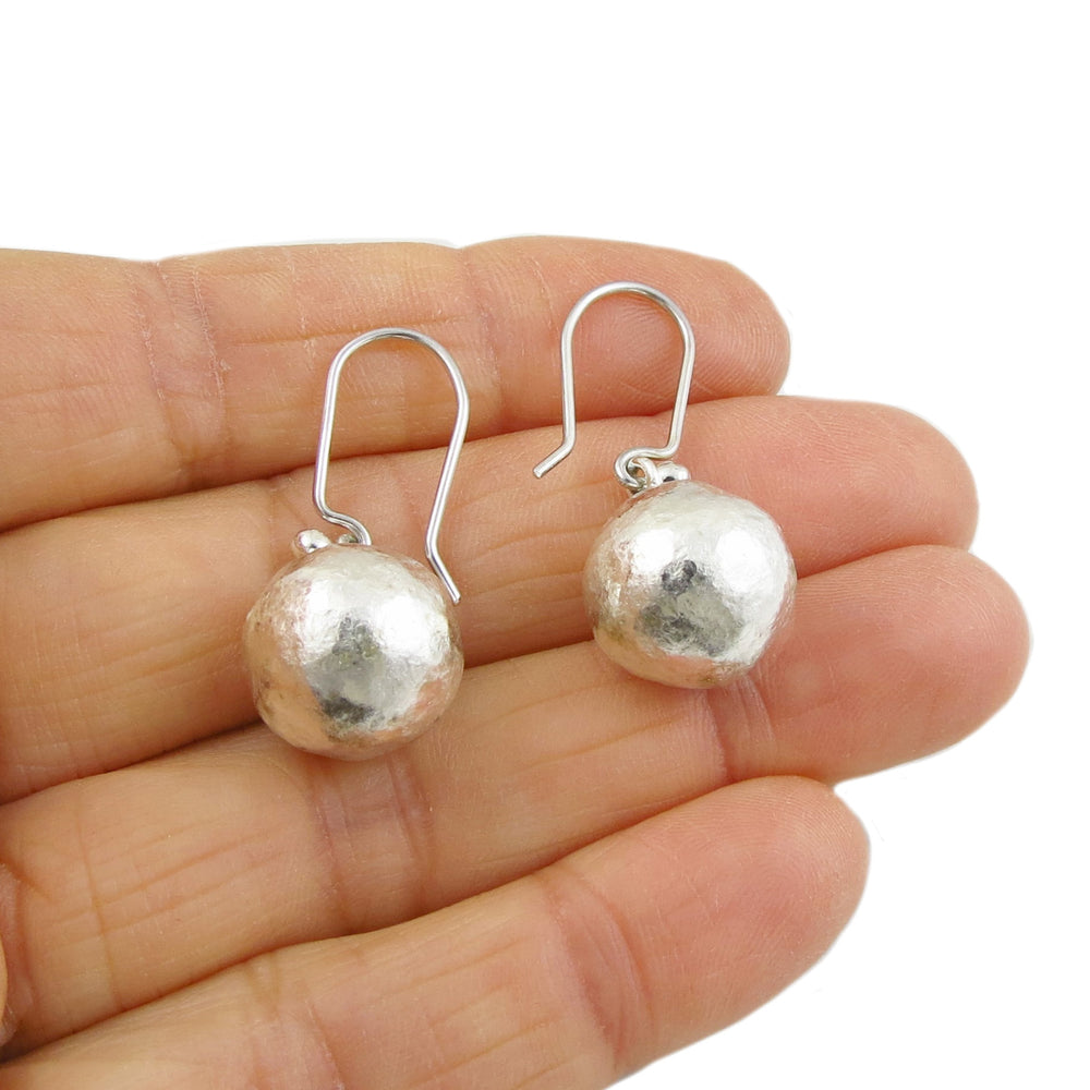 A pair of battered silver ball bead earrings held in a person's hand, with a matte finish and long hook fittings.