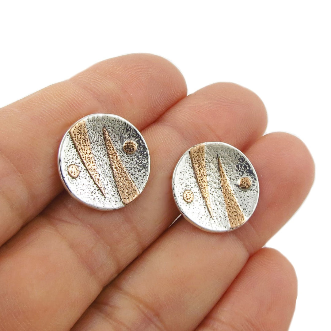 A pair of round mixed metal 925 silver stud earrings with textured metallic copper accents, held on a person's fingers against a white background.