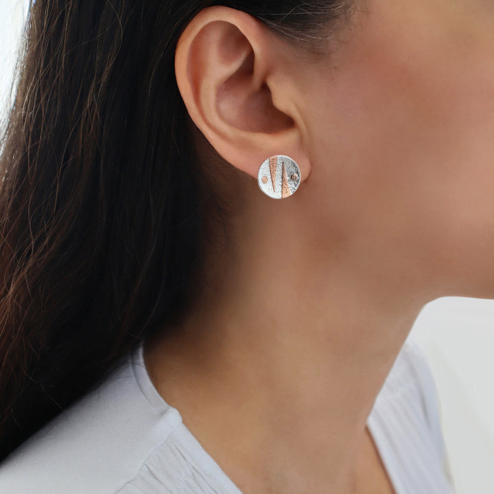 A close-up of a woman's ear wearing a small round mixed metal 925 silver and copper stud earring with a textured, geometric pattern of copper inclusions. 
