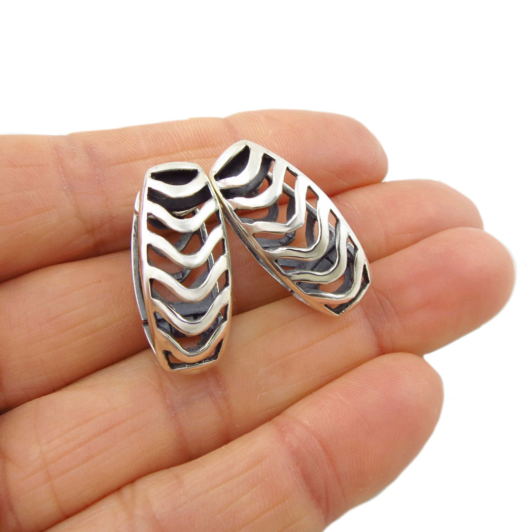 A pair of large sterling silver huggie earrings with a silhouette pattern held in a person's hand against a white background.