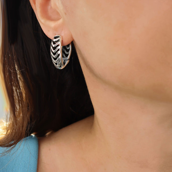 A close-up of a woman wearing a silver huggie hoop earring with a black and white wavy pattern, shown against skin and dark hair.