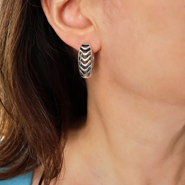 A close-up of a woman's ear wearing large front and back sterling 925 silver huggies hoop earring with a textured chevron pattern.