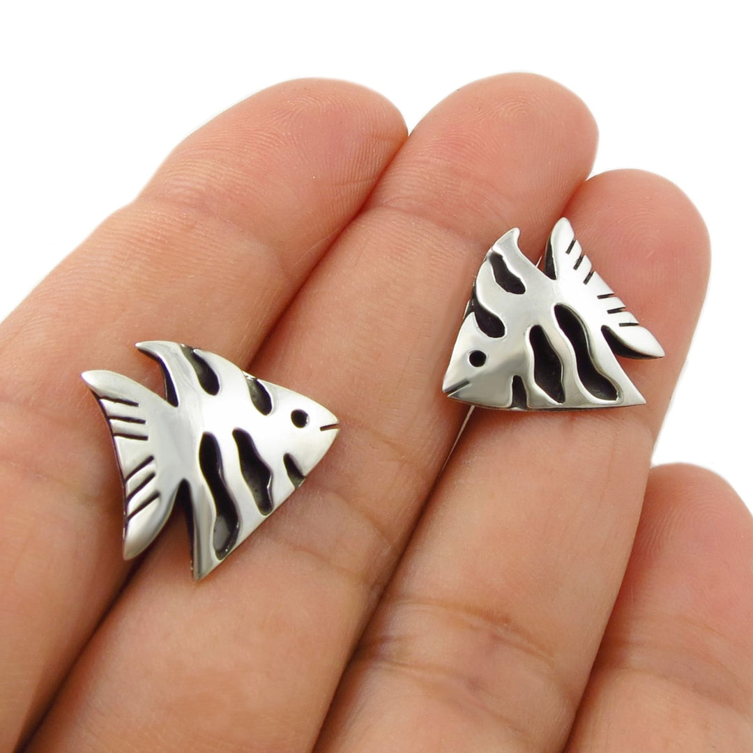 A pair of Mexican 925 silver fish-shaped stud earrings with black shadow box details, shown resting on a woman's fingers against a white background.
