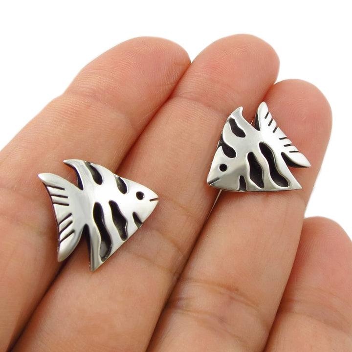 A pair of Mexican 925 silver fish-shaped stud earrings with black shadow box details, shown resting on a woman's fingers against a white background.