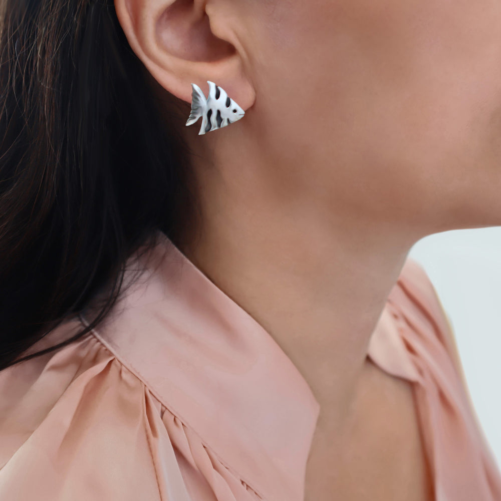 A close-up of a woman's ear wearing a small sterling 925 silver angel fish stud earring, with dark hair and a peach-coloured blouse visible against a light background.