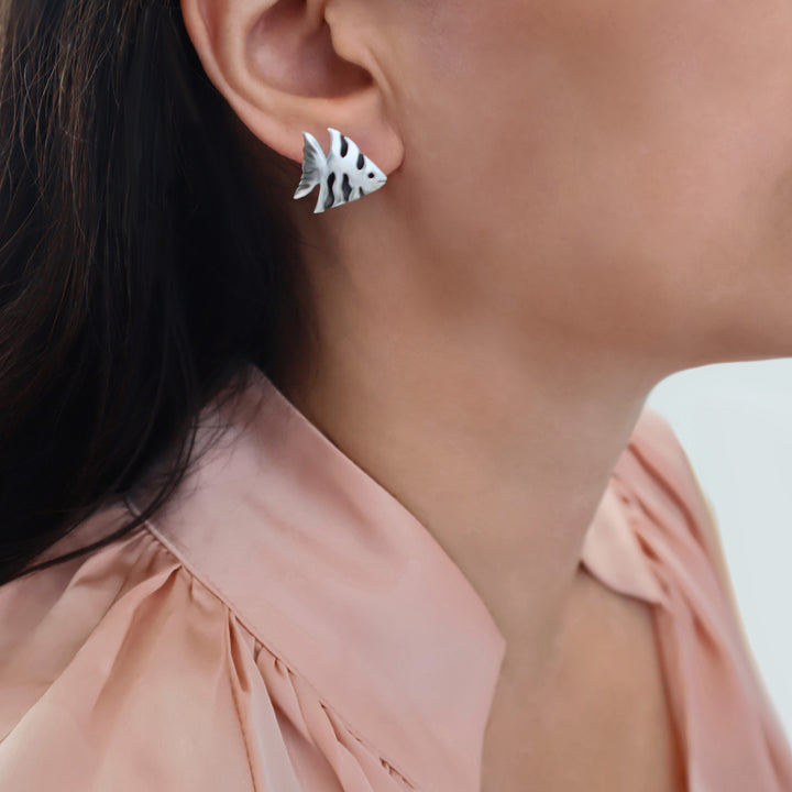 A close-up of a woman's ear wearing a small sterling 925 silver angel fish stud earring, with dark hair and a peach-coloured blouse visible against a light background.