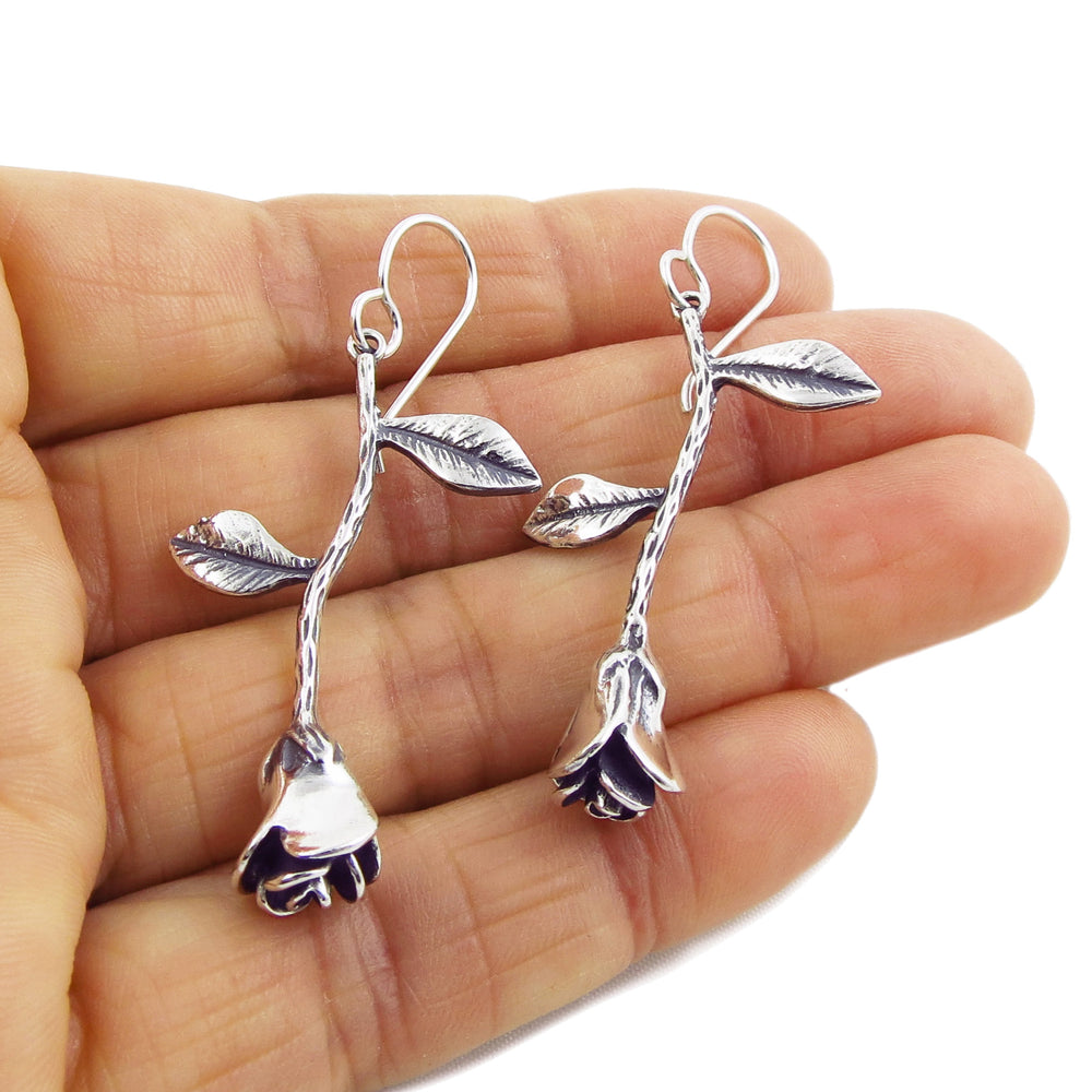 A pair of silver drop earrings designed with rosebud motifs, held in a hand against a white background.