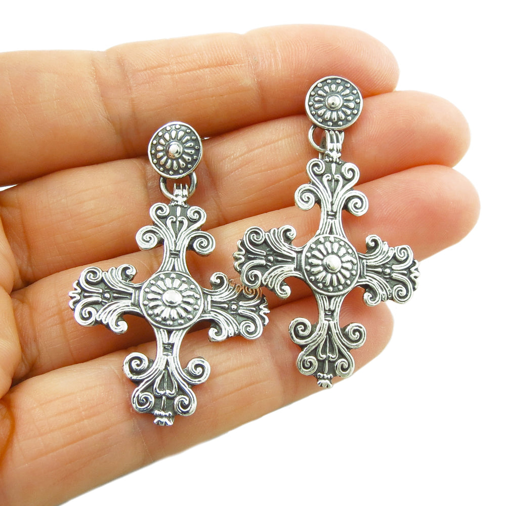 A pair of baroque style Mexican Taxco 925 sterling silver earrings with ornate cross designs, held in a hand.