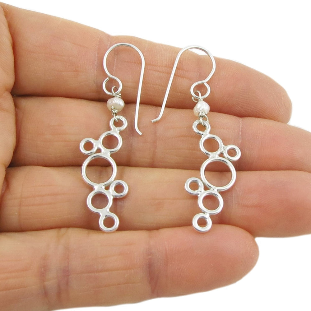 A pair of silver bubble earrings with cluster design and freshwater pearls, hanging on hooks, displayed on a hand