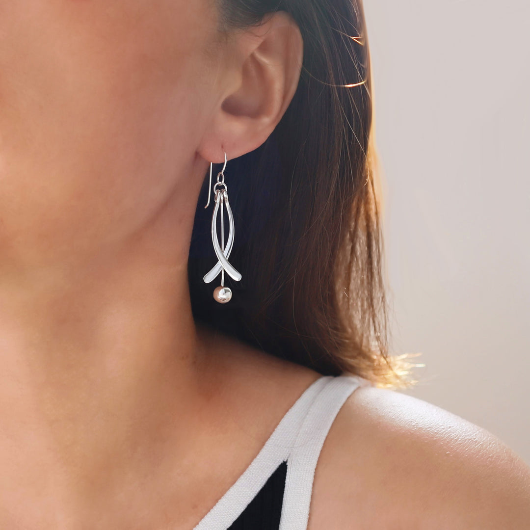 Close-up of a woman wearing a long silver earring decorated with a dangling ball bead on a neutral background