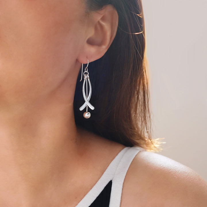 Close-up of a woman wearing a long silver earring decorated with a dangling ball bead on a neutral background