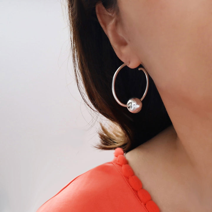 Close-up of a woman wearing a silver hoop earring with a large round hammered ball bead accent on a plain background.