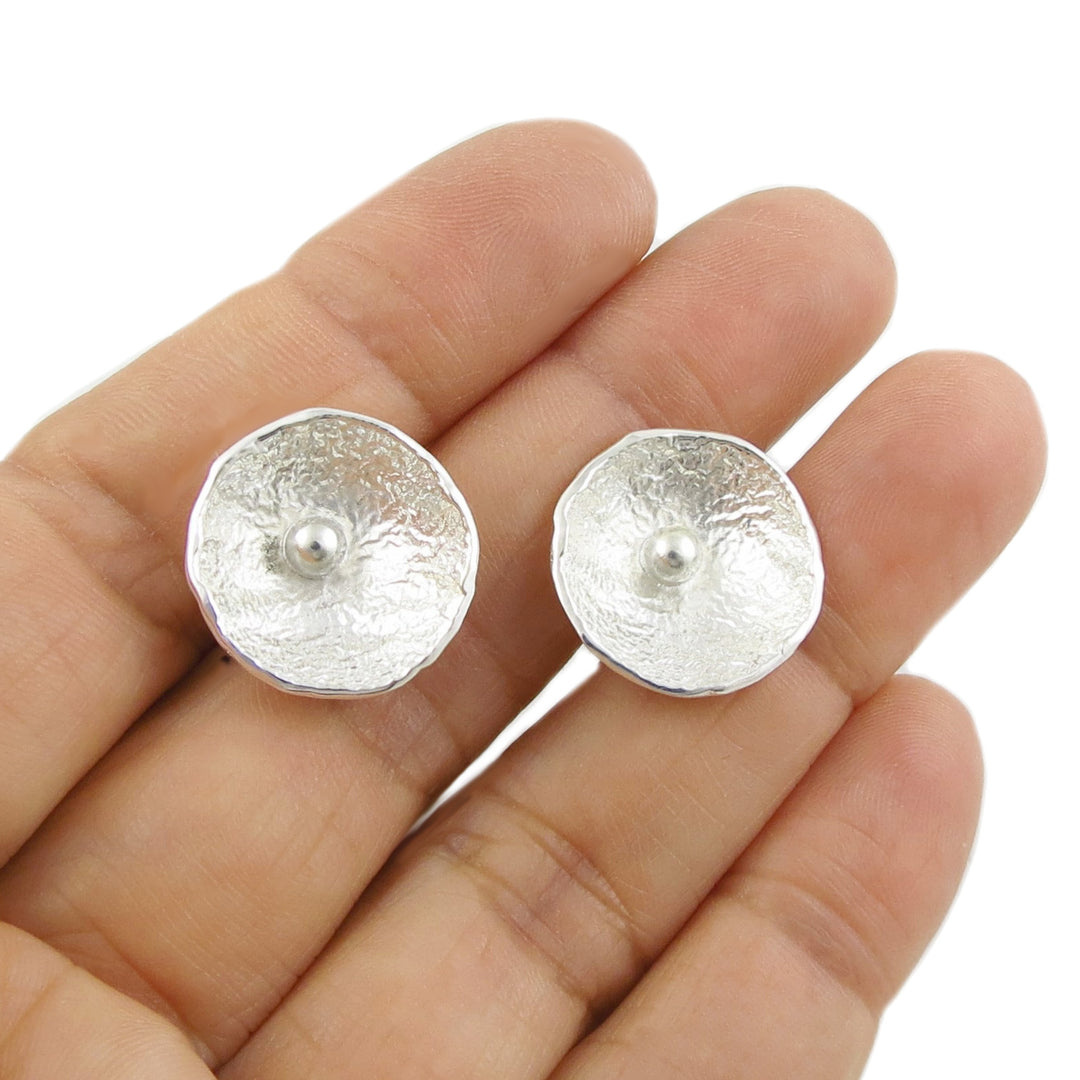 Silver poppy flower stud earrings held between fingers on a white background. 