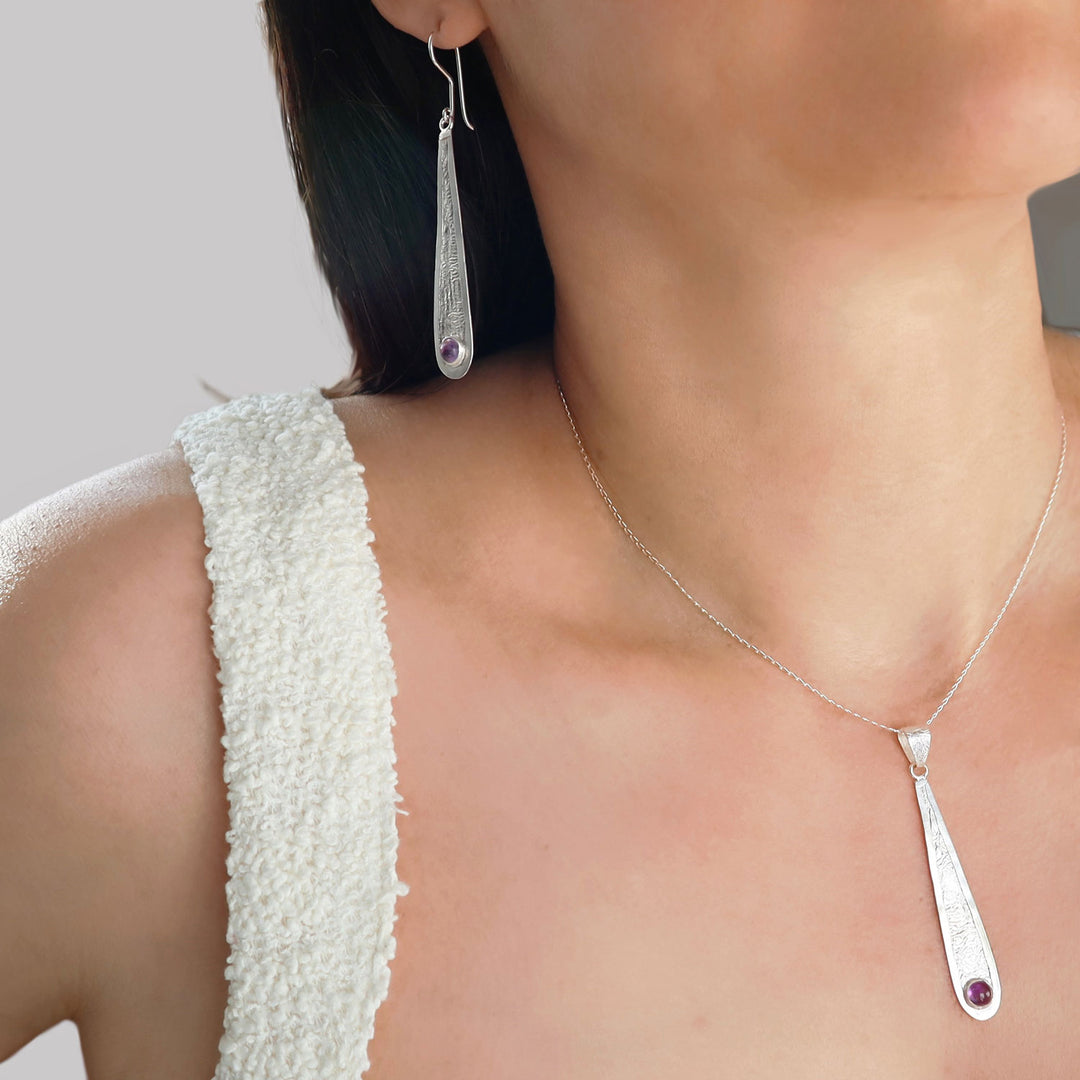 Silver teardrop pendant necklace and matching drop earring worn by a model, featuring a small purple amethyst gemstone accent at the bottom of each piece.