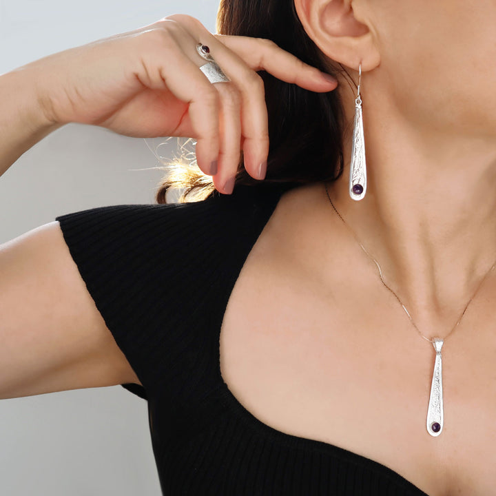 A close-up lifestyle shot of a model wearing a matching silver-toned jewelry set, featuring a teardrop-shaped drop earring and a delicate necklace with a matching pendant, both adorned with small dark gemstone accents at the bottom.