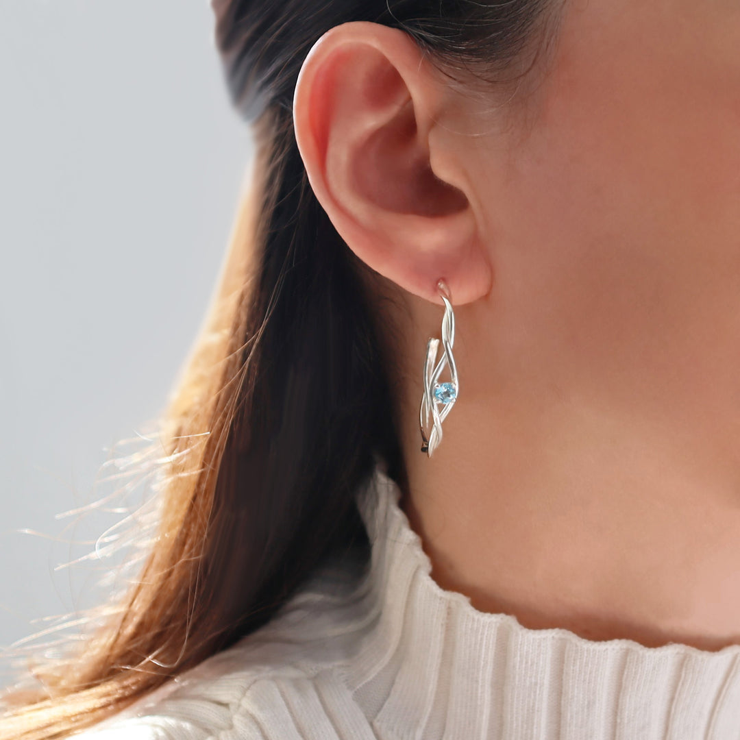 Close-up of a person wearing a silver earring with a blue topaz gemstone against a neutral background