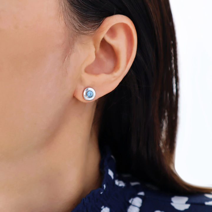 Close-up of an ear wearing a silver earring with a faceted blue topaz gemstone on a white background