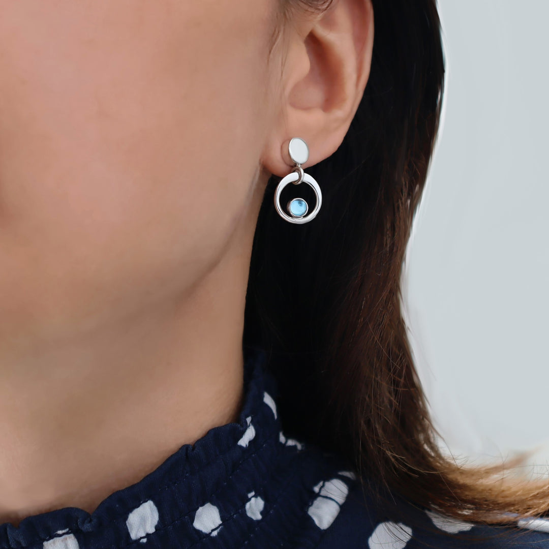 Close-up of a person wearing a silver earring with a blue topaz gemstone on a neutral background