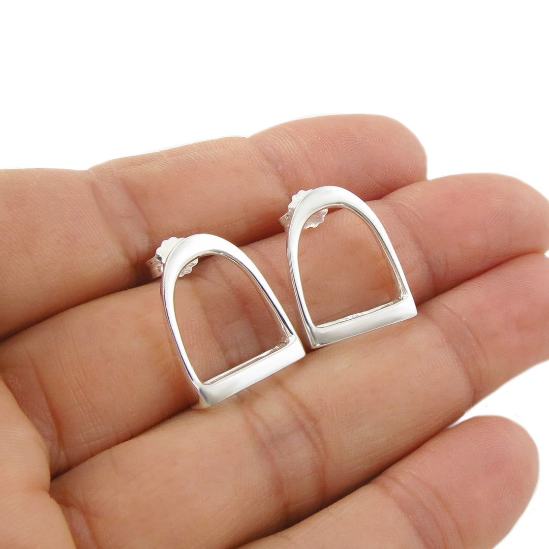 A pair of large 925 sterling silver equestrian-style horse stirrup earrings held between fingers on a white background