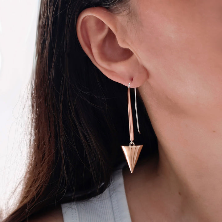 A close-up of a woman wearing a long, thin mixed metal copper and 925 silver dangle earring with a triangular cone-shaped drop, shown against skin and dark hair.