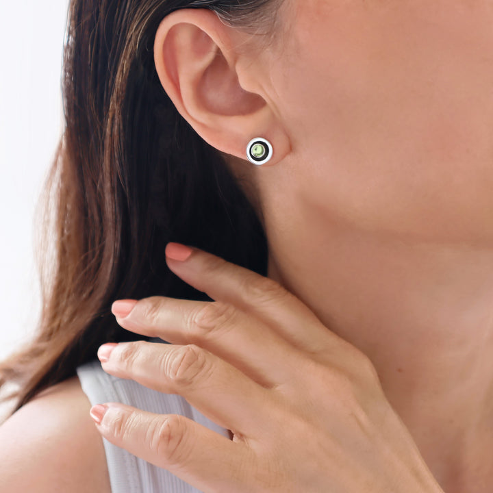 A close-up of a woman's ear wearing a small round stud earring with a white outer ring and a green peridot gemstone in the middle, with her hand near her neck.
