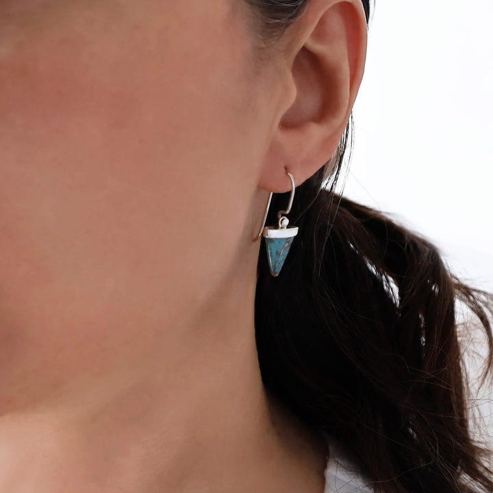 A close-up side profile of a woman wearing a silver earring with a large triangular turquoise stone.