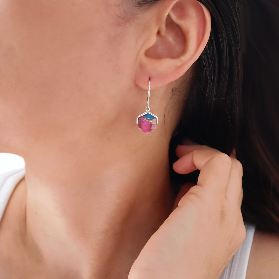 A close-up of a female model wearing a silver geometric drop earring with a faceted pink purple spiney gemstone. 