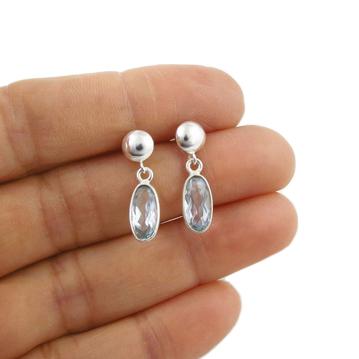 A pair of silver dangle earrings with faceted oval cut blue topaz gemstones, held between a woman's fingers against a white background.