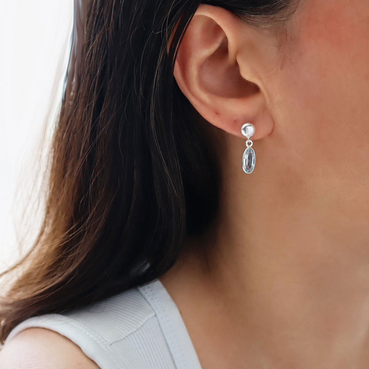 A close-up of a woman's ear wearing a silver drop earring with a brilliant cut faceted blue topaz  gemstone. 