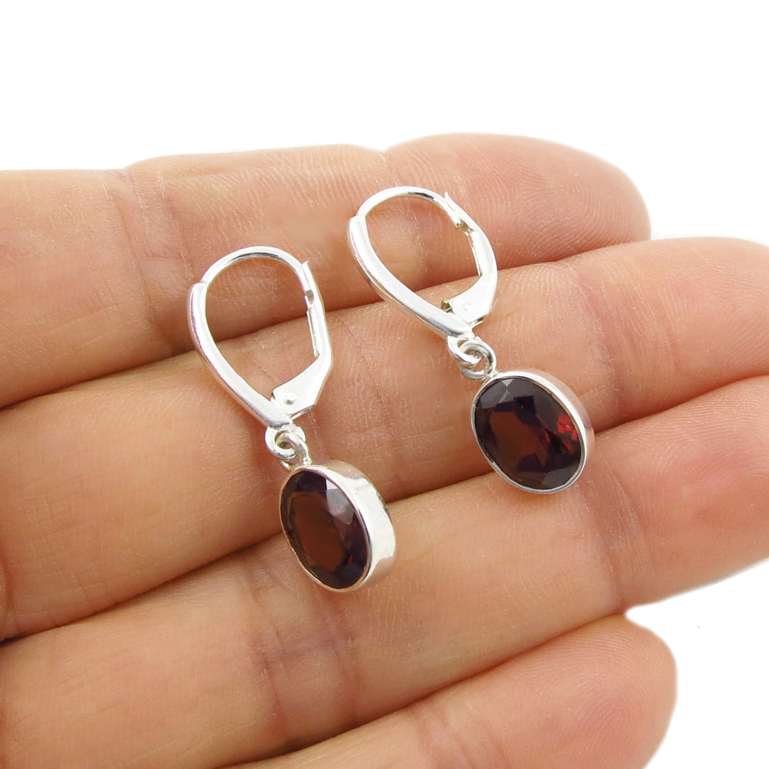 A pair of deep red faceted garnet January birthstone 925 sterling silver earrings with lever-back fasteners shown in a person's hand on a white background. 