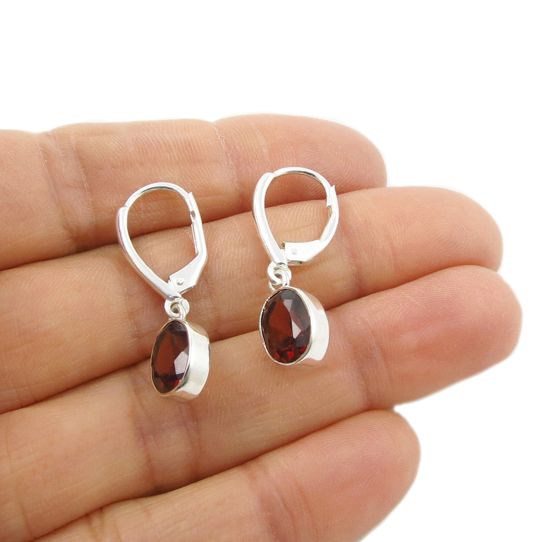 A pair of silver lever-back earrings with faceted dark red garnet gemstone drops, shown resting on a person's hand against a white background.